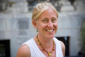 Photo of Dr. Eva Purkey who is smiling in front of a faded backdrop of a building. She is wearing a white sleeveless shirt and necklace with blonde hair. 