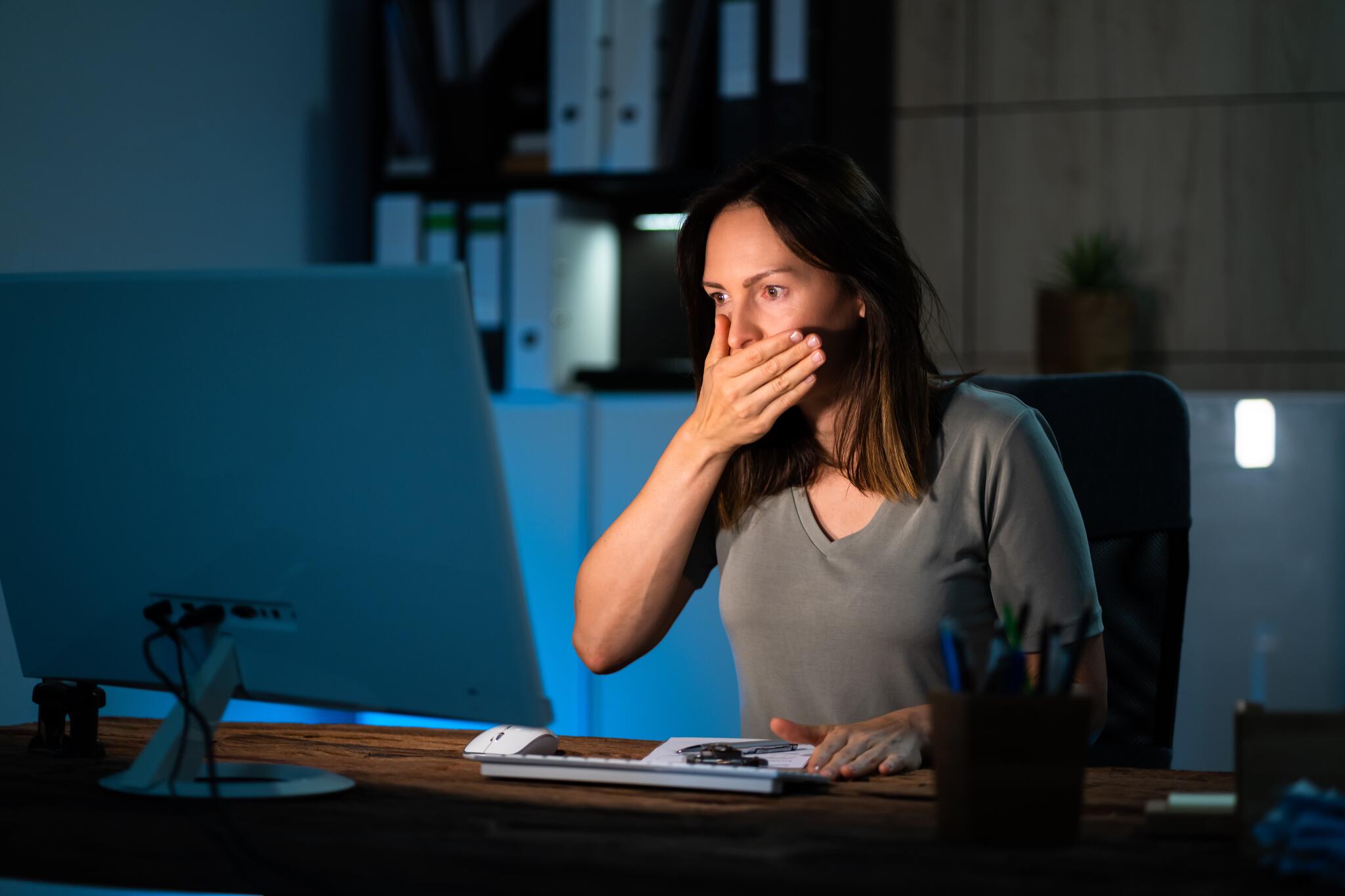 A woman sits in front of a computer with a shocked expression on her face. Her hand covers her mouth.