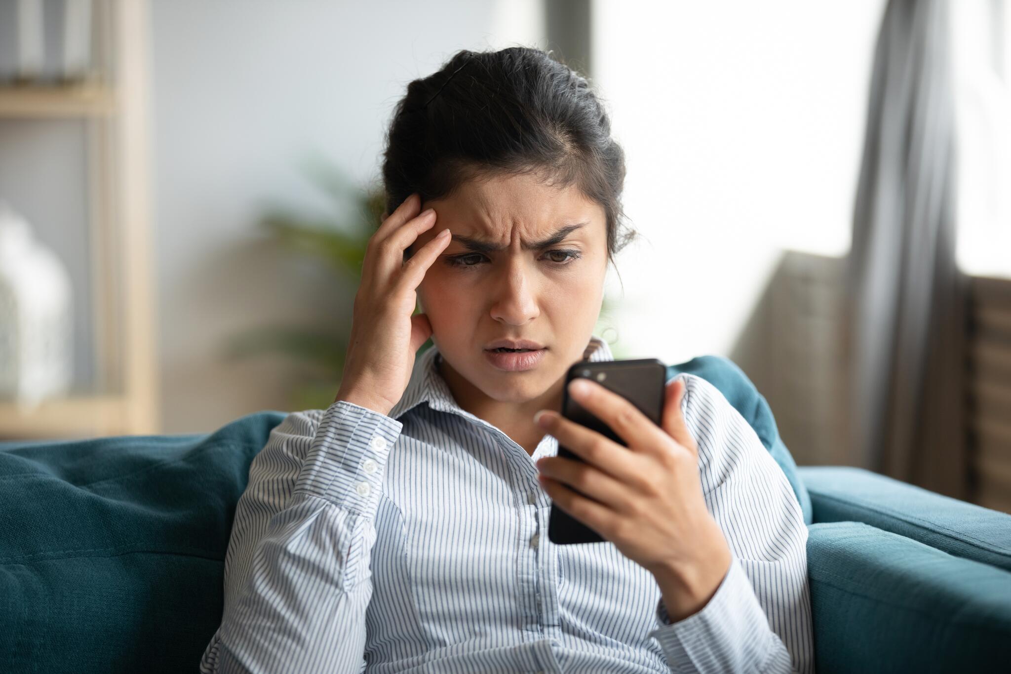 A woman sits on a couch and stares in horror at her phone.