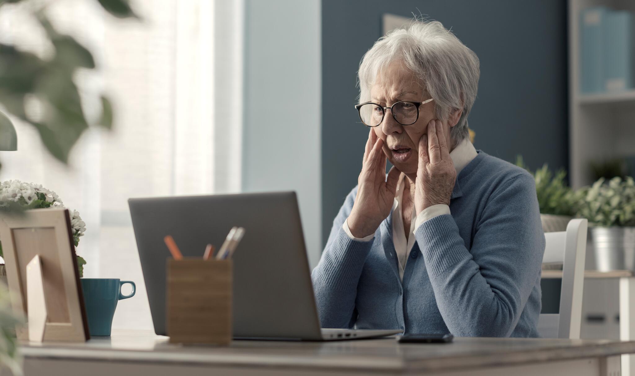An elderly woman sits in front of a laptop. Her hands frame her face in a look of shock as she gazes at her laptop screen.