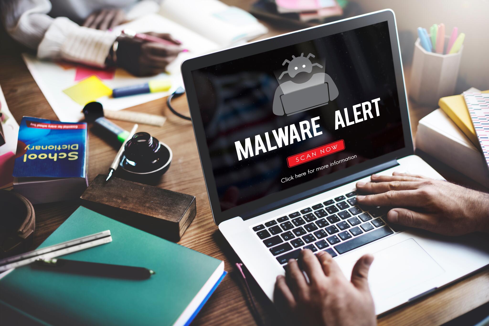 A man sits in front of a laptop. An image of a virus with the word "malware" appears on his laptop screen.