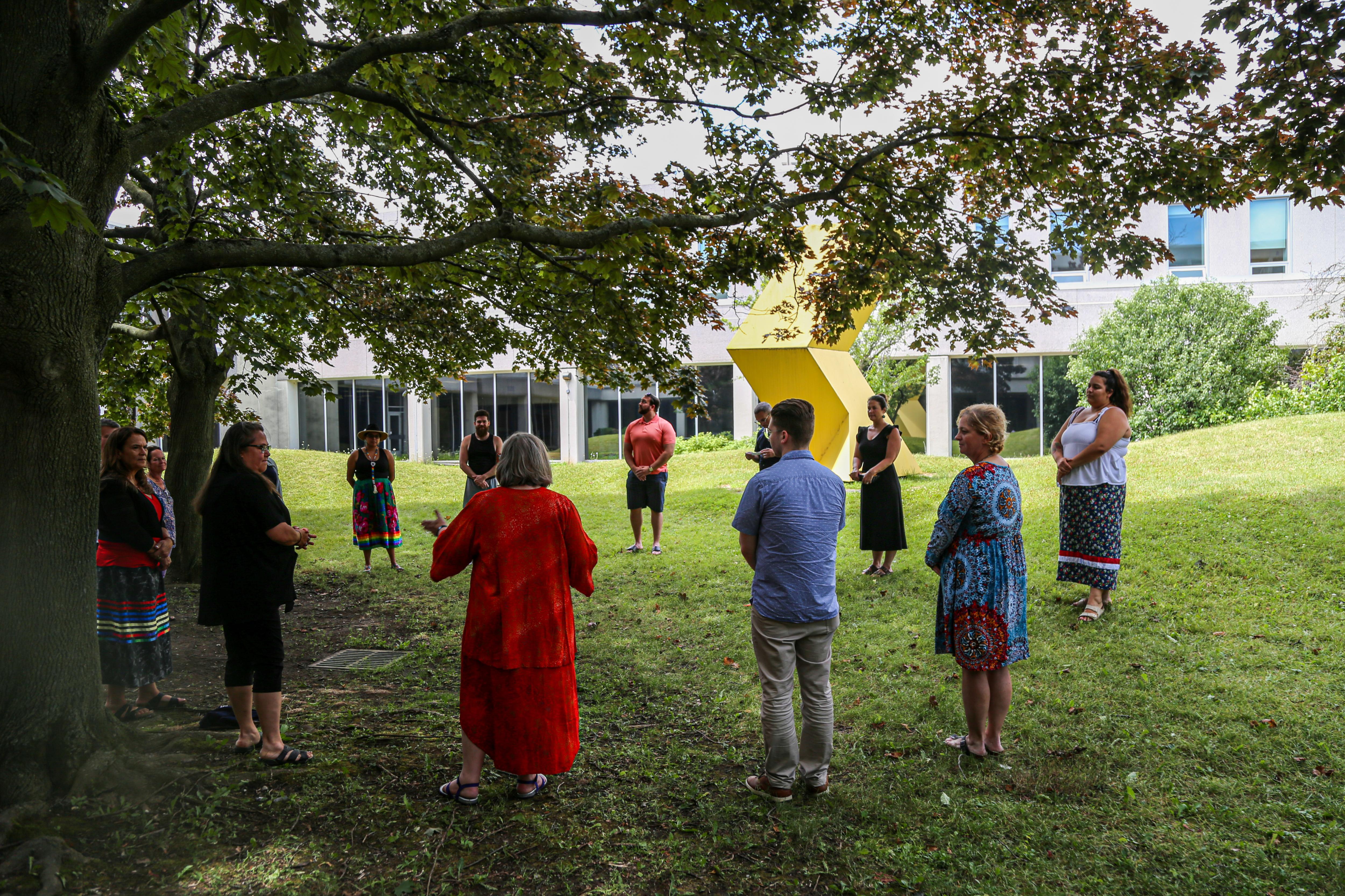A class of wise students stands in a circle outside with a tree over them 