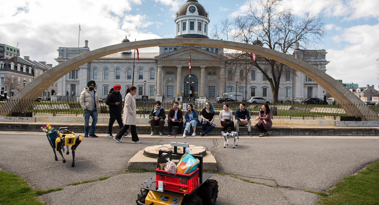 3 students standing and 7 sitting in front of a fountain with 3 robots in front of them