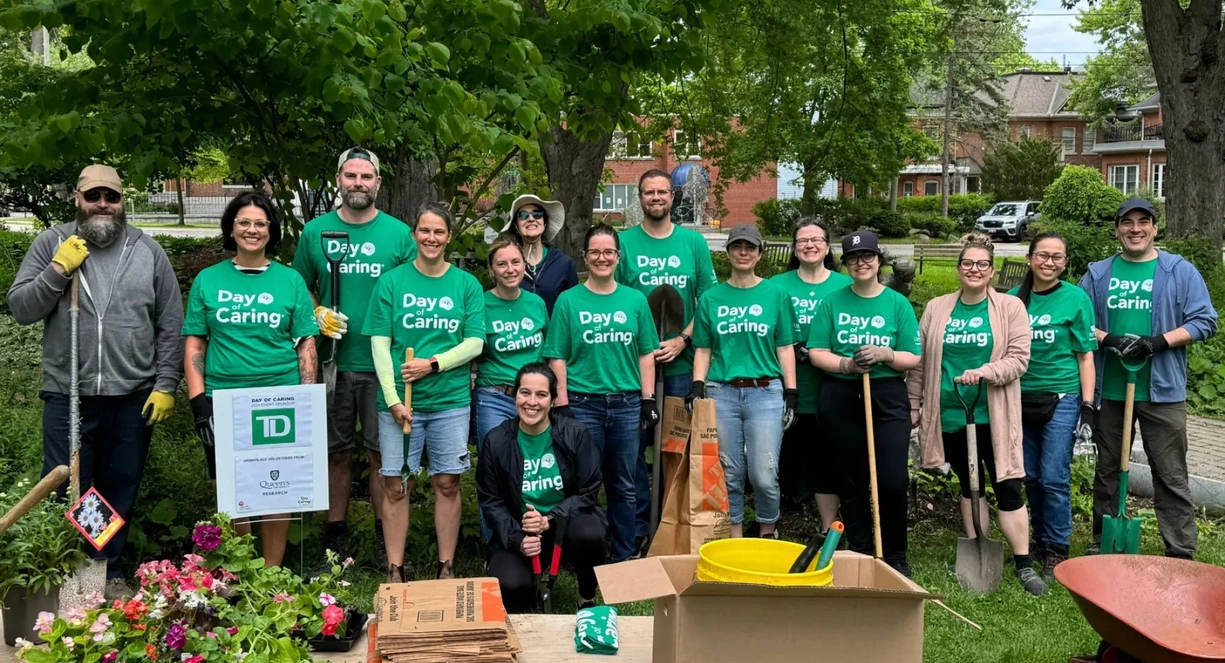 15 people in green t-shirts standing with shovels and rakes in a garden